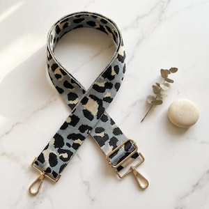 May include: A wide, adjustable bag strap featuring a woven leopard print pattern in shades of light blue, cream, and black. The strap includes gold-toned metal hardware, specifically two swivel lobster clasps and a rectangular sliding buckle.