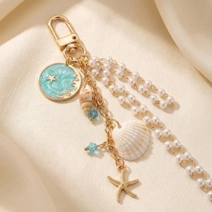 Op de afbeelding: Een goudkleurige sleutelhanger met een ronde bedel met lichtblauwe emaille afwerking en een gouden ster, een kleine spiraalschelp, een witte mantelschelp, een goudkleurige zeesterbedel, twee kleine blauwe kralen en twee strengen witte kunstparels.