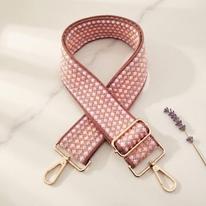 May include: A woven bag strap featuring a repeating diamond pattern in shades of light pink and white. The strap is finished with gold-toned metal hardware, including two swivel lobster clasps and an adjustable sliding buckle for length control.
