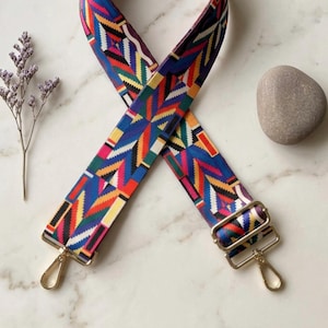 May include: A wide, adjustable bag strap featuring a vibrant geometric pattern in shades of blue, red, green, yellow, and white. The strap includes polished gold-tone metal hardware, including a sliding buckle and two swivel lobster claw clasps.
