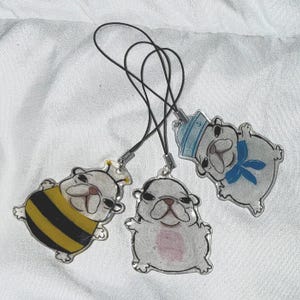 May include: Three clear acrylic bulldog charms. One is dressed as a bee with yellow and black stripes, another has a pink belly, and the third wears a sailor hat and blue bow. Each charm has a black cord attached.