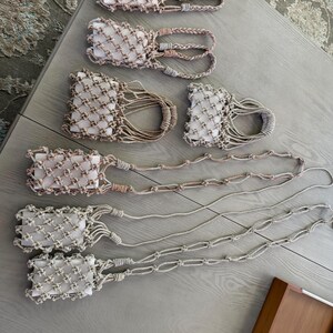 May include: A collection of seven rectangular white soap bars encased in beige and light pink macrame cord holders. Four holders feature short braided handles, while three holders are attached to long, knotted macrame cord straps for hanging.