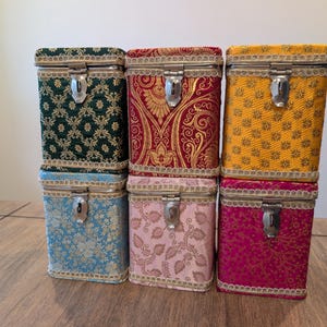 Op de afbeelding: Zes vintage dozen met stoffen bekleding in verschillende kleuren, waaronder groen, rood, geel, blauw, roze en magenta. Elke doos heeft een decoratief patroon, een metalen sluiting en sierlijsten. De dozen zijn gestapeld op een houten oppervlak.