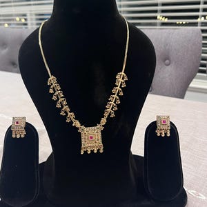 Puede incluir: Un conjunto de collar y pendientes dorados exhibido sobre un busto negro. El collar presenta un colgante cuadrado con una piedra roja y detalles intrincados. Los pendientes a juego también tienen piedras rojas.