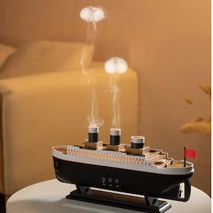 Puede incluir: Un difusor de aceites esenciales con forma de barco en blanco y negro con tres chimeneas y una bandera roja. El difusor emite vapor blanco, creando un efecto de medusa. El producto está sobre una mesa blanca.