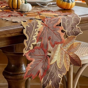 May include: Embroidered leaf-shaped table runner in shades of brown, orange, and gold. The runner is 175cm long and 12.7cm wide, with detailed stitching. Pumpkins and a cake are on the table, suggesting a fall theme.