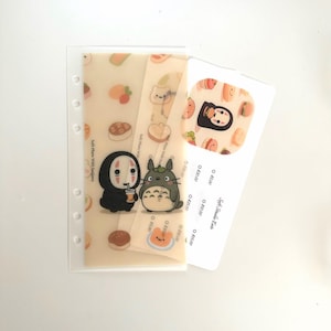 May include: A clear plastic planner insert with illustrations of food and anime characters. The insert features images of a character from Spirited Away, Totoro, and various food items. A white card with text is also included.