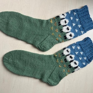 Könnte beinhalten: Ein Paar handgestrickte Socken. Die Socken sind hauptsächlich grün, mit blauen Bündchen und einem Muster aus weißen Schafen mit schwarzen Gesichtern und kleinen gelben Akzenten. Aus weichem, strukturiertem Garn.