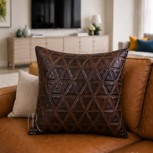 May include: A square dark brown leather throw pillow featuring a geometric pattern of stitched triangles across the front surface. The pillow includes a small logo in the bottom corner with the text ARSAM RUGS. It is placed on a tan leather sofa.