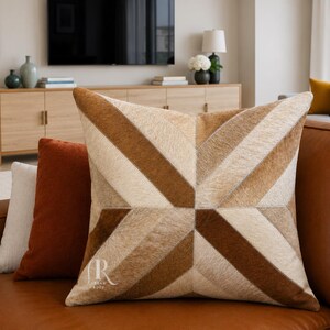 May include: A square throw pillow featuring a geometric pattern made of stitched panels of hair-on hide in shades of cream, tan, and dark brown. The design consists of diagonal stripes radiating from the center to form a star-like motif.