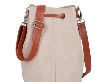 May include: A beige bucket bag with brown leather straps and accents. The bag has a drawstring closure and silver-colored metal hardware. The adjustable shoulder strap is attached with metal clasps. The bag is made of a textured fabric.