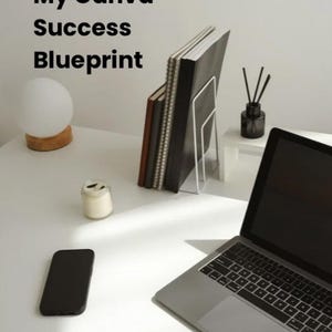 May include: A laptop, notebooks, and a smartphone are on a white desk. The text "From Creativity to Freedom" and "My Canva Success Blueprint" are visible. A small lamp and candle are also present, suggesting a workspace.