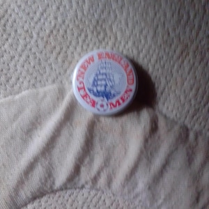 Op de afbeelding: Een ronde, witte knoop met rode tekst "NEW ENGLAND TEA MEN" rond een blauwe illustratie van een zeilschip. De knoop ligt op een getextureerd, gebroken witte ondergrond.