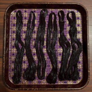 May include: Six bundles of black embroidery floss are arranged on a decorative tray. The tray has a purple and gold patterned fabric lining. The floss is neatly arranged, showcasing its texture and color.