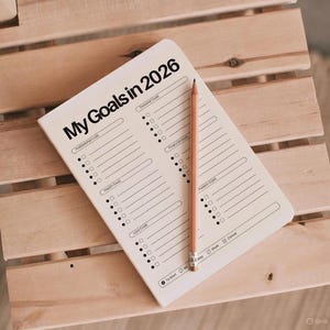 May include: A cream-colored notebook with the text "My Goals in 2026" and a wooden pencil. The notebook has sections for goals, including professional, personal, and financial goals. The notebook is on a light brown wooden surface.