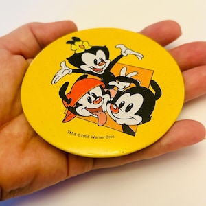 Puede incluir: Un botón amarillo con los personajes de dibujos animados de Warner Bros., Yakko, Wakko y Dot, con el texto "TM & ©1995 Warner Bros." El botón tiene un fondo amarillo con los tres personajes de dibujos animados en negro, blanco y rojo.