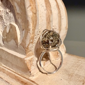 May include: A silver-toned ring featuring a cluster of pyrite stone, wire-wrapped in a cage-like design. The ring has an adjustable band. The pyrite stone has a metallic, gold-like appearance. The ring is set against a light-colored, textured wooden surface.