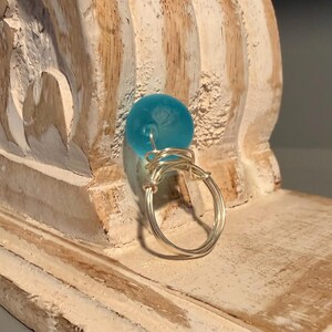 May include: A handmade ring featuring a round, light blue bead set atop a silver wire band. The wire is intricately wrapped around the bead and forms the ring's circular shape. The ring is displayed on a distressed, light-colored wooden surface.