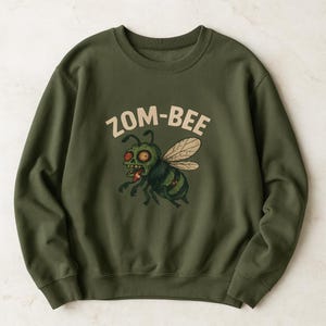 May include: Olive green sweatshirt with the text "ZOM-BEE" above a cartoon zombie bee illustration. The bee has red eyes, green skin, and a striped abdomen. The design is centered on the front of the long-sleeved crew neck.