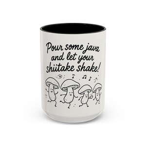 May include: White ceramic mug with a black interior. The mug has the text "Pour some java and let your shiitake shake!" and dancing mushroom illustrations. A fun, whimsical design for tea or coffee.