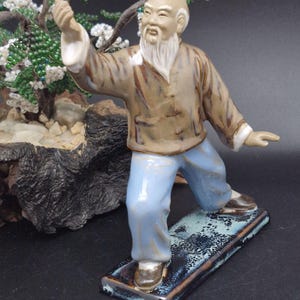 Może przedstawiać: Ceramiczna figurka starszego mężczyzny w pozie sztuk walki. Postać nosi brązową kurtkę, jasnoniebieskie spodnie i brązowe buty. Figurka stoi na prostokątnej podstawie z niebiesko-czarną glazurą. Małe drzewko bonsai w tle.