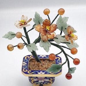 May include: A decorative bonsai tree sculpture with orange and white flowers, orange berries, and green leaves. The tree is planted in a blue and gold enamel pot. The sculpture is set against a white background.