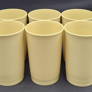 May include: Six cream-coloured, cylindrical cups are arranged in two rows on a dark surface. The cups have a smooth, matte finish and a simple, rounded rim. They are evenly spaced, suggesting a set.