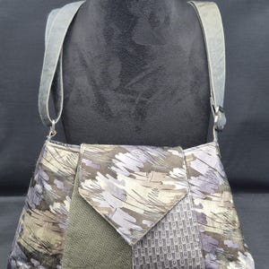 May include: A patchwork shoulder bag with a grey suede strap. The bag features a variety of fabrics in shades of grey, lavender, and olive green. The flap is a geometric shape. The bag is displayed on a black mannequin.