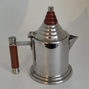 Peut inclure: Une cafetière en acier inoxydable poli avec un corps cylindrique, un couvercle articulé surmonté d'un cône à gradins et une poignée latérale dotée d'une prise en bois brun. La cafetière présente un petit bec triangulaire et une base circulaire.