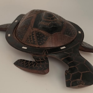 May include: A dark brown wooden turtle-shaped box with a removable lid. The shell features intricate geometric carvings and small white rectangular inlays along the rim. The flippers and head are detailed with etched patterns resembling scales.