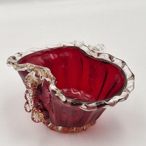 May include: A small, heart-shaped glass bowl in a deep red colour. The rim is clear with a ruffled edge, and the base has a gold-coloured trim. The side of the bowl has a decorative gold-coloured embellishment. The bowl is likely for decorative purposes.