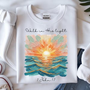 May include: White sweatshirt with a colorful sunset design. The artwork features an orange sun over blue and teal waves, with the text "Walk in His light" and "1 John 1:7". The sweatshirt is made of heavy cotton.