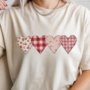 Puede incluir: Camiseta color crema con cuatro apliques en forma de corazón. Cada corazón tiene un patrón único: floral, cuadros rojos, pequeñas flores rosas y cuadros rojos y blancos. Los corazones están delineados con costuras negras, creando un aspecto encantador y artesanal.