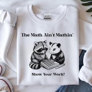 May include: White sweatshirt with the text "The Math Ain't Mathin'" above an illustration of a raccoon and a possum working on a calculator. The text "Show Your Work!" is below the illustration.