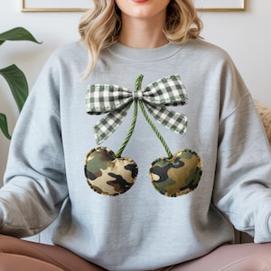 May include: A pale grey sweatshirt with a playful cherry design. The cherries are made from camouflage fabric, with green stems and a gingham bow. The design is centrally placed on the chest.