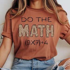Può includere: Una maglietta color ruggine con la scritta "DO THE MATH" in nero. La parola "MATH" è riempita con un motivo leopardato. Sotto il testo c'è l'equazione (9X7)+4.