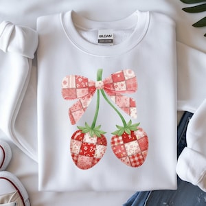 May include: White sweatshirt featuring a watercolor design of two strawberries with a bow. The strawberries and bow are a patchwork of red and pink patterns. The sweatshirt has a crew neck and long sleeves.