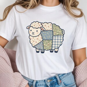 May include: White t-shirt with a cartoon sheep design. The sheep is made of patchwork fabric in floral, blue, and gingham patterns. The sheep has a cream-coloured face and a fluffy head. The design is outlined in black.
