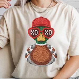 May include: Cream-colored t-shirt featuring a whimsical duck design. The duck wears a red cap and heart-shaped sunglasses with "XO" on each lens. The duck has a green head, brown body, and a yellow beak. The design is centered on the shirt.