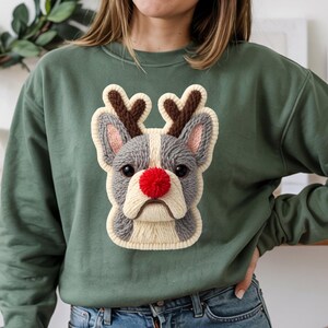 Puede incluir: Sudadera verde con un diseño texturizado de un bulldog francés con cuernos de reno y una nariz roja. La cara del perro es gris, blanca y beige, con cuernos marrones y orejas rosas. Una prenda festiva con temática navideña.