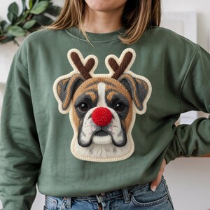May include: Green sweatshirt featuring a Boxer dog with reindeer antlers and a red nose. The dog's face is detailed with brown, white, and black accents, and the antlers are brown. The design is a festive, holiday-themed graphic.