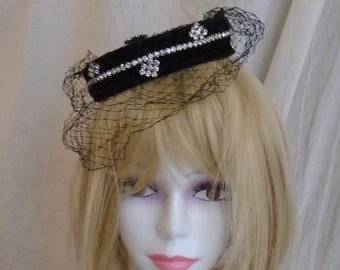 May include: A black velvet hat with a rhinestone trim and a black net veil. The hat is worn on a white mannequin head with blonde hair.