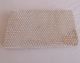 May include: A rectangular, silver-toned clutch covered in small, sparkling rhinestones. The clutch has a slightly rounded shape and a textured surface, reflecting light. The overall design is elegant and suitable for formal events.