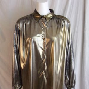May include: A metallic gold button-down shirt with long sleeves. The shirt has a classic collar and buttoned cuffs. The fabric has a shiny, reflective quality, giving it a glamorous appearance. The shirt is displayed on a mannequin.