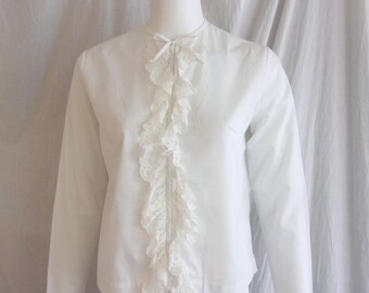 May include: A white long-sleeved blouse with a high collar and a delicate lace ruffle down the front. The cuffs also feature lace trim. A small bow detail is at the neckline. The blouse is displayed on a mannequin.