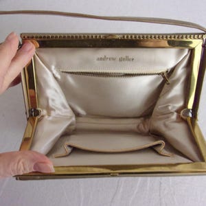 May include: Open vintage handbag with a gold-toned frame and a beige handle. The interior features a beige satin lining, a zippered pocket, and the text "andrew geller". The bag is open, revealing its interior.