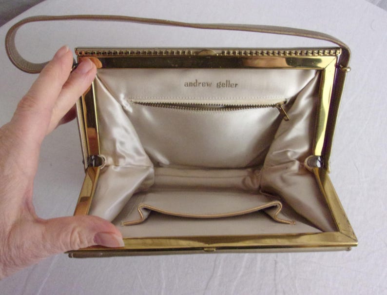 May include: Open vintage handbag with a gold-toned frame and a beige handle. The interior features a beige satin lining, a zippered pocket, and the text "andrew geller". The bag is open, revealing its interior.