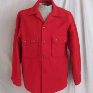 May include: A bright red wool shirt with two chest pockets and button closures. The long-sleeved shirt has a collared neckline and is displayed on a mannequin. The shirt is a solid color.