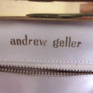 May include: Close-up of a white fabric with the embossed text "andrew geller" in a gold color. A gold-colored metal frame is visible at the top, and a zipper is at the bottom. The image suggests a handbag or purse.