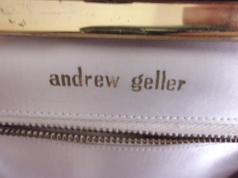 May include: Close-up of a white fabric with the embossed text "andrew geller" in a gold color. A gold-colored metal frame is visible at the top, and a zipper is at the bottom. The image suggests a handbag or purse.